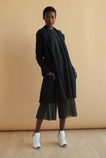 An adult person with a deep skin tone models a long black jacket, jumpsuit, and white heels. 