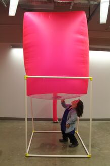 An adult person with a fair skin tone, medium-length brown hair, and dwarfism, stands under an installation by Bri Beck.