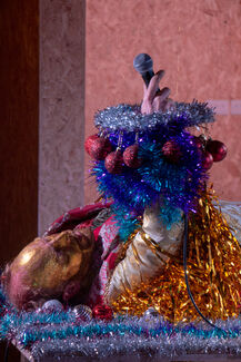 A performer dressed in glitter and tinsel lies on their back, holding a microphone.