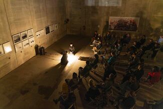 Two people sit on the floor with a light between them and a seated audience observing. 