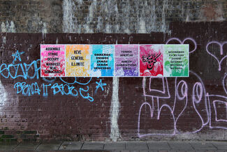 Wheat paste installation by Aaron Hughes, reading "ASSEMBLE, STRIKE, OCCUPY, MANIFEST REAL DEMOCRACY," featuring colorful panels in various languages. 