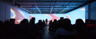 A person performing against a lit background, with audience visible in the foreground.