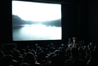 A theater full of people watching a projection of a body of water and two people playing instruments on stage. 