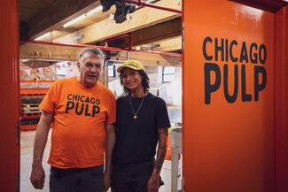 Two adult people of varying ages, genders and skintones standing next to an orange wall that reads "CHICAGO PULP."