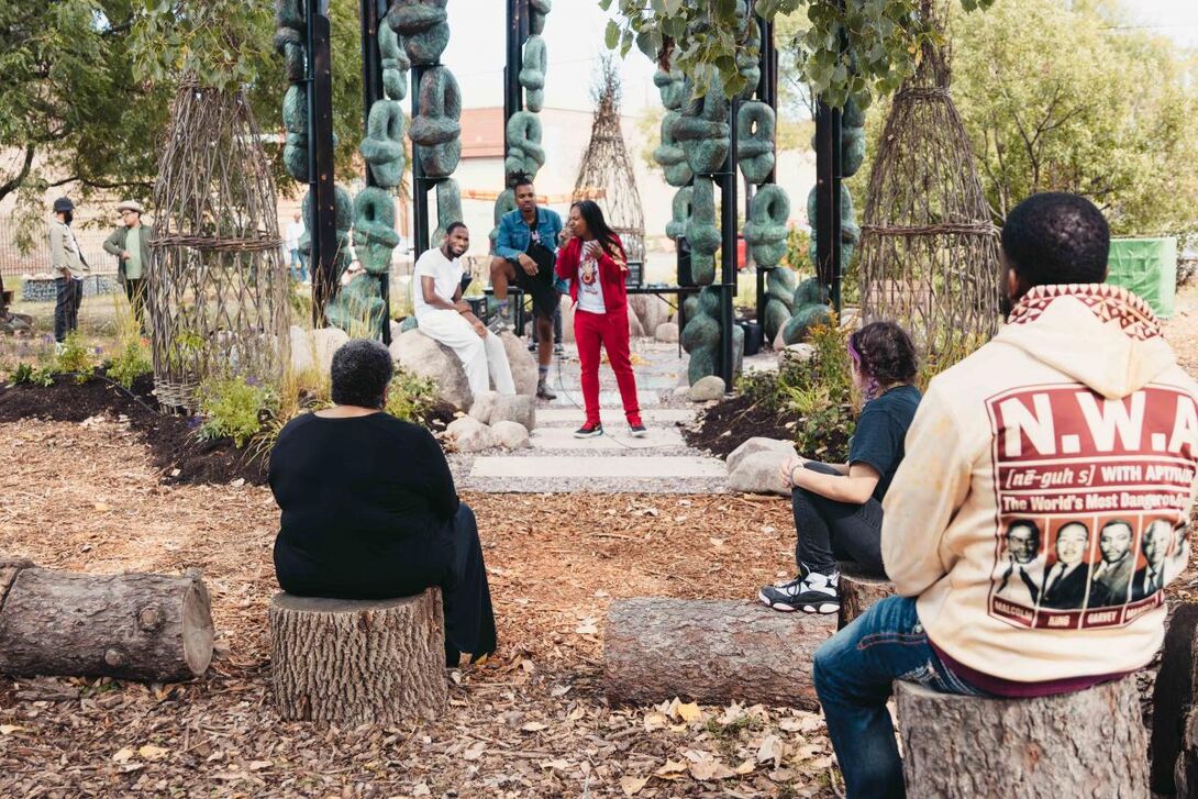 Group of people seated outside on tree stumps listening to a speaker standing under a gazebo-like structure surrounded by sculptural objects.