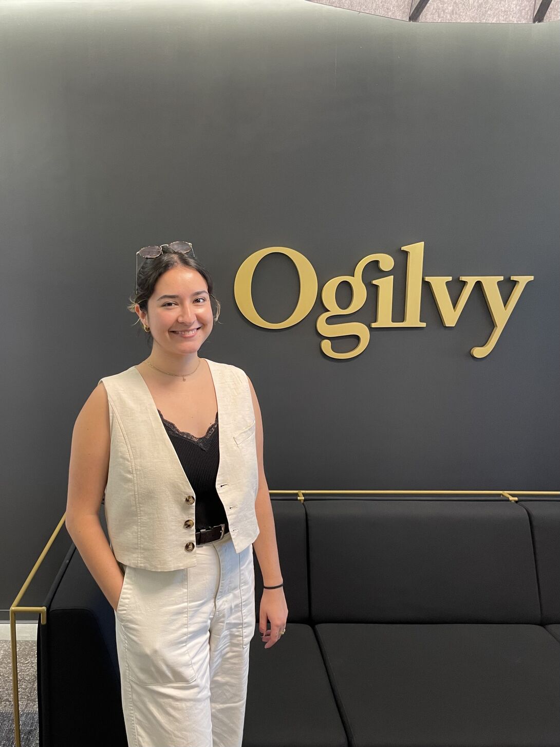 A woman stands wearing a cropped white vest and white pants poses in front of a sign that says "Ogilvy"