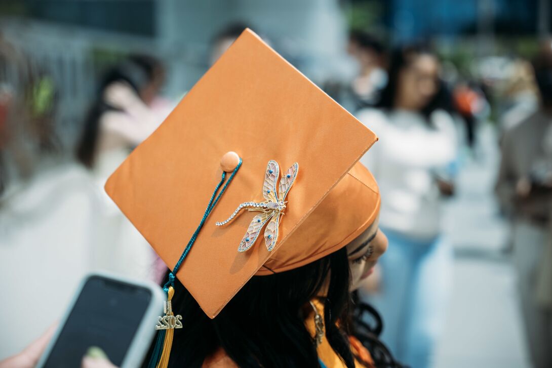 A student in an orange graduation cap with a dragonfly brooch.