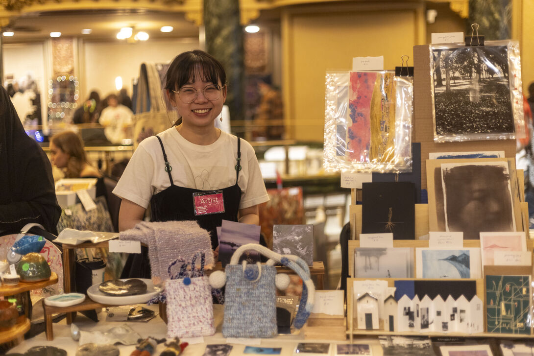 An Asian person wearing an "artist" badge smiling and standing behind a display of various artworks. 