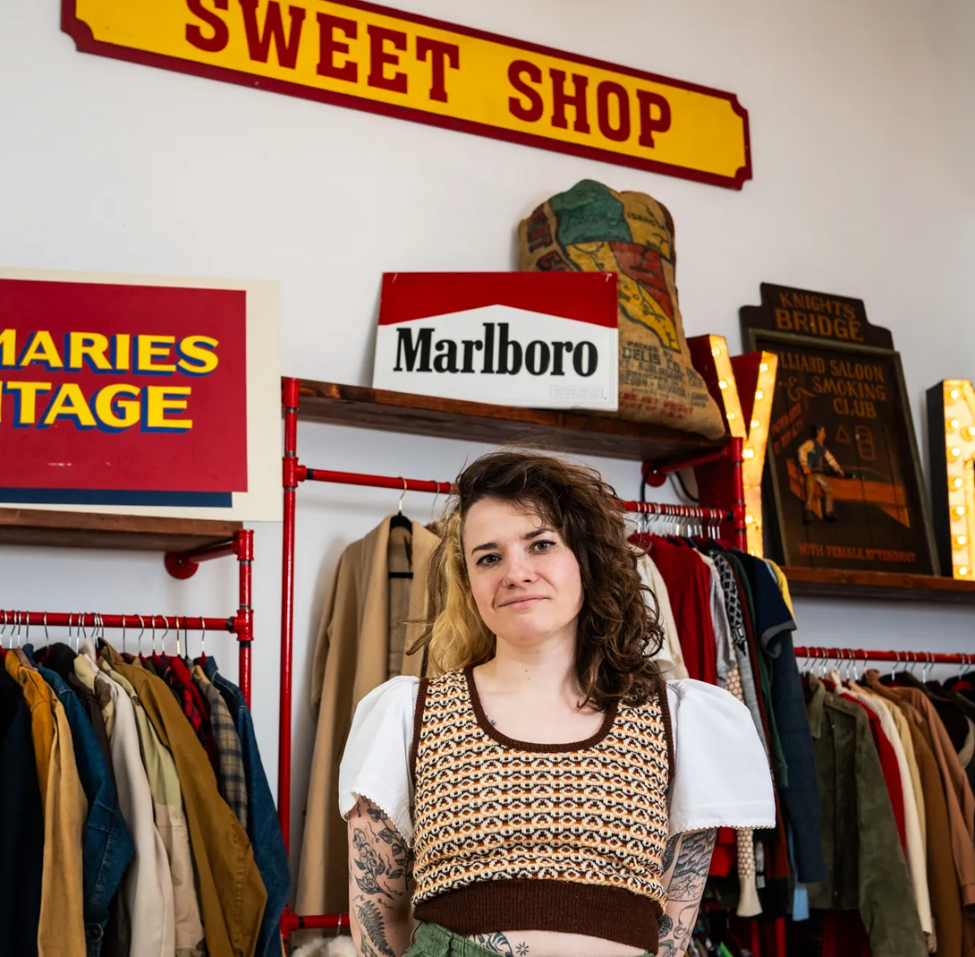 Person standing in a vintage clothing store beneath a “Sweet Shop” sign, surrounded by racks of colorful clothes and retro signage.