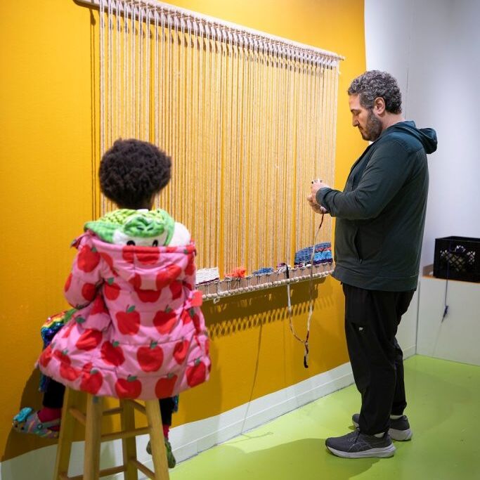 Child and adult working together on a large vertical weaving loom mounted to a yellow wall. The child sits on a stool wearing a bright red apple-patterned coat, while the adult stands, threading yarn into the loom.