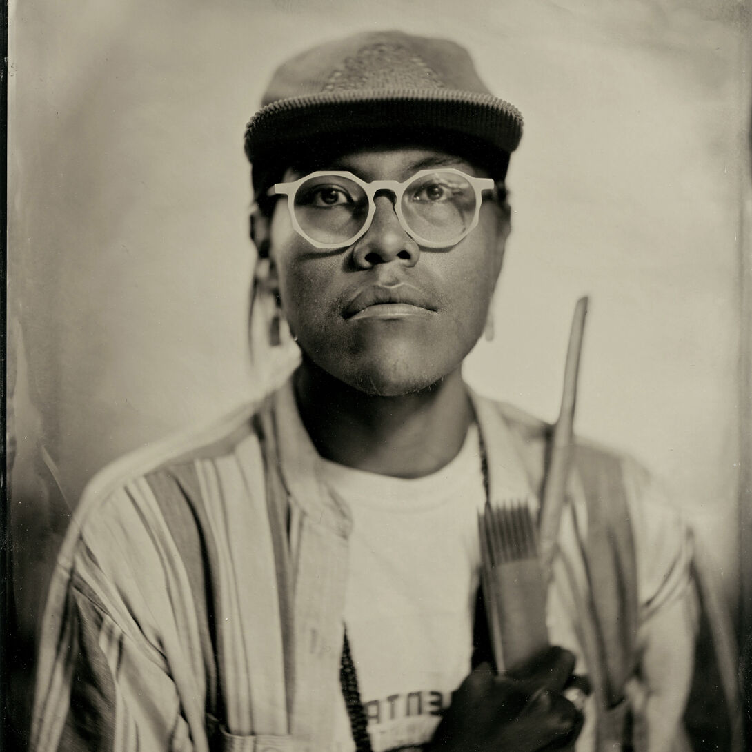 TinType headshot of Tyrell Tapaha holding Diné weaving tools over their heart.