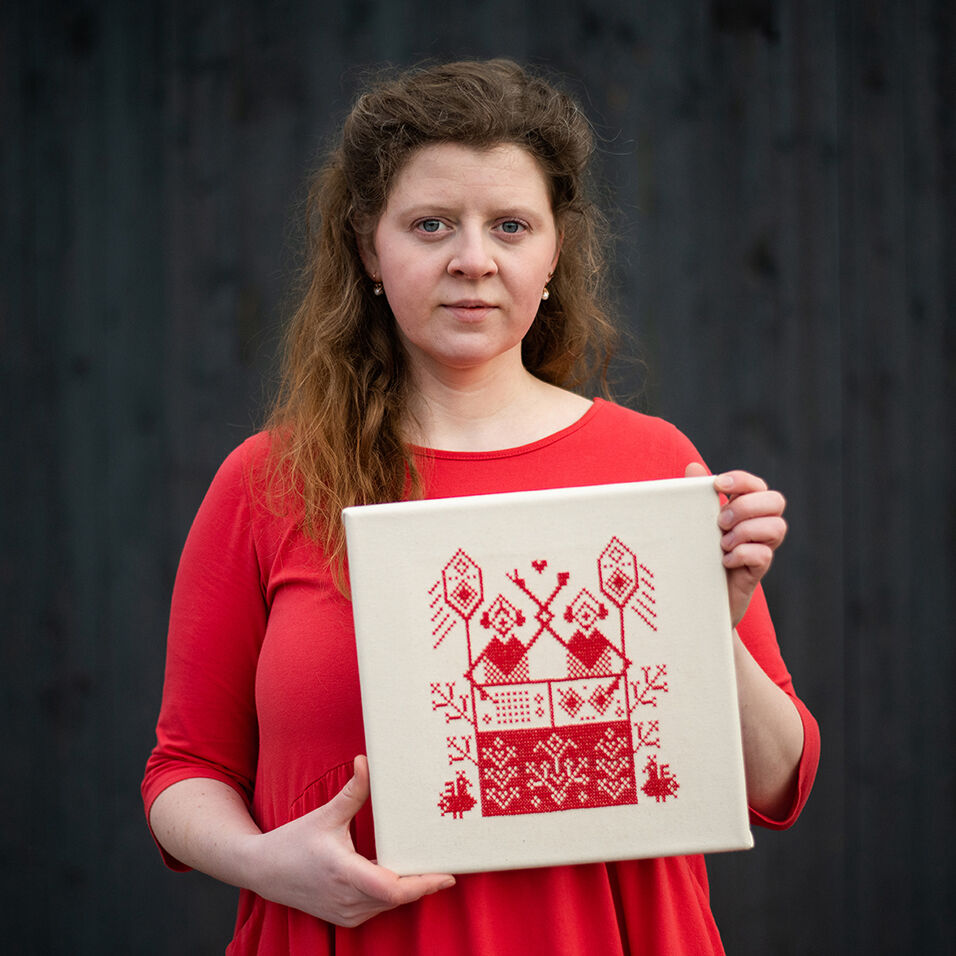 The artist in a red dress, holds a cream-colored square with red embroidery.