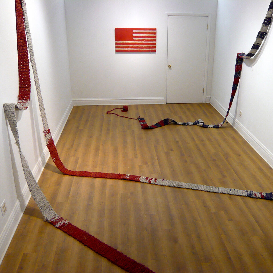 Excessively long, red white and blue crocheted scarves cascade from  white walls and criss-cross the wooden floor in a small exhibition space. On the back wall is an American flag in red and white.