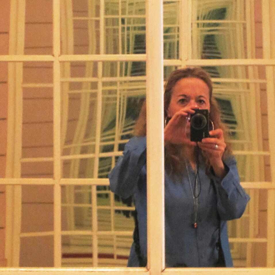 The artist holds a vertical camera blocking her nose and mouth in front of a paned window, which echoes to infinity in the background.