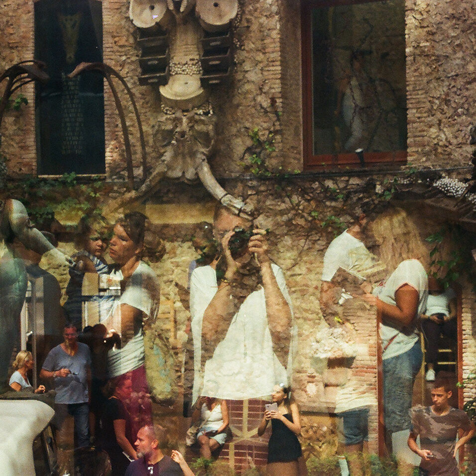 Grainy, double-exposed photo with the artist in the center holding a camera to her eye. She is surrounded by other human figures interacting in front of a stone building.