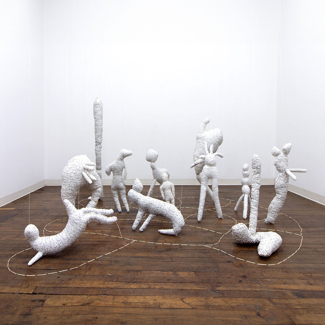 Knee-high white sculptures of human and animal figures sit on a wooden floor in a white-walled gallery space. A dashed white line weaves between and around them on the floor. 