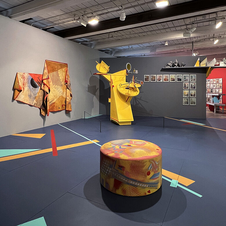 Ottoman-like sculpture covered in printed textile with lines, triangles and circles in primary colors in the foreground of a museum space with light grey walls and a dark grey floor. In the background is a cascading textile with similar patterning hung on the wall and a larger than human-sized, angular, yellow sculpture.