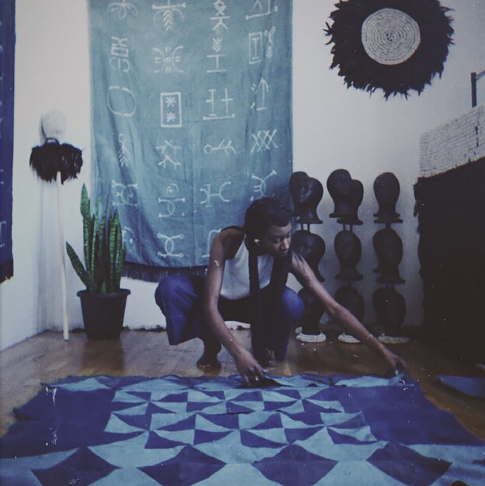 The artist squats to arrange a pieced textile in a variety of blues. Behind her, hangs a indigo-dyed textile and other sculptures. 