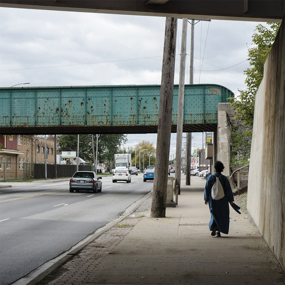 The artist wears a backpack and walks away from the camera down a city sidewalk.