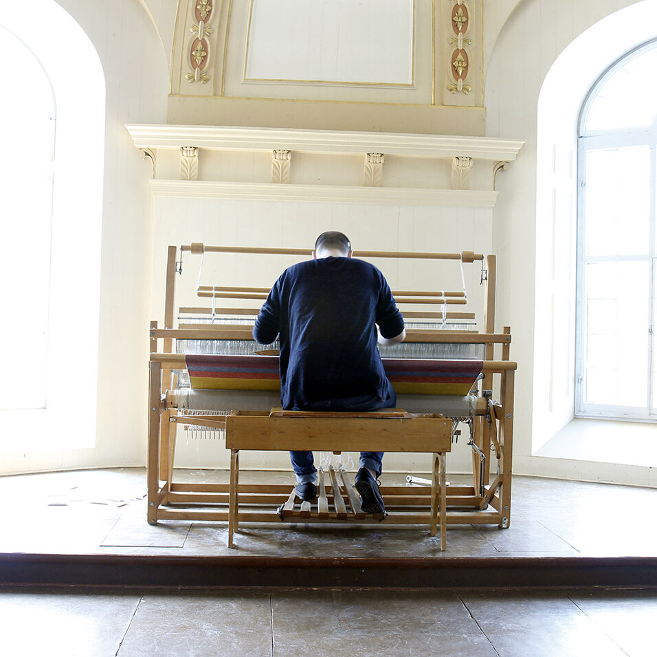 The artist, dressed all in black and flanked by two large windows, is hunched over a floor loom, evoking the posture of an organist in a church.