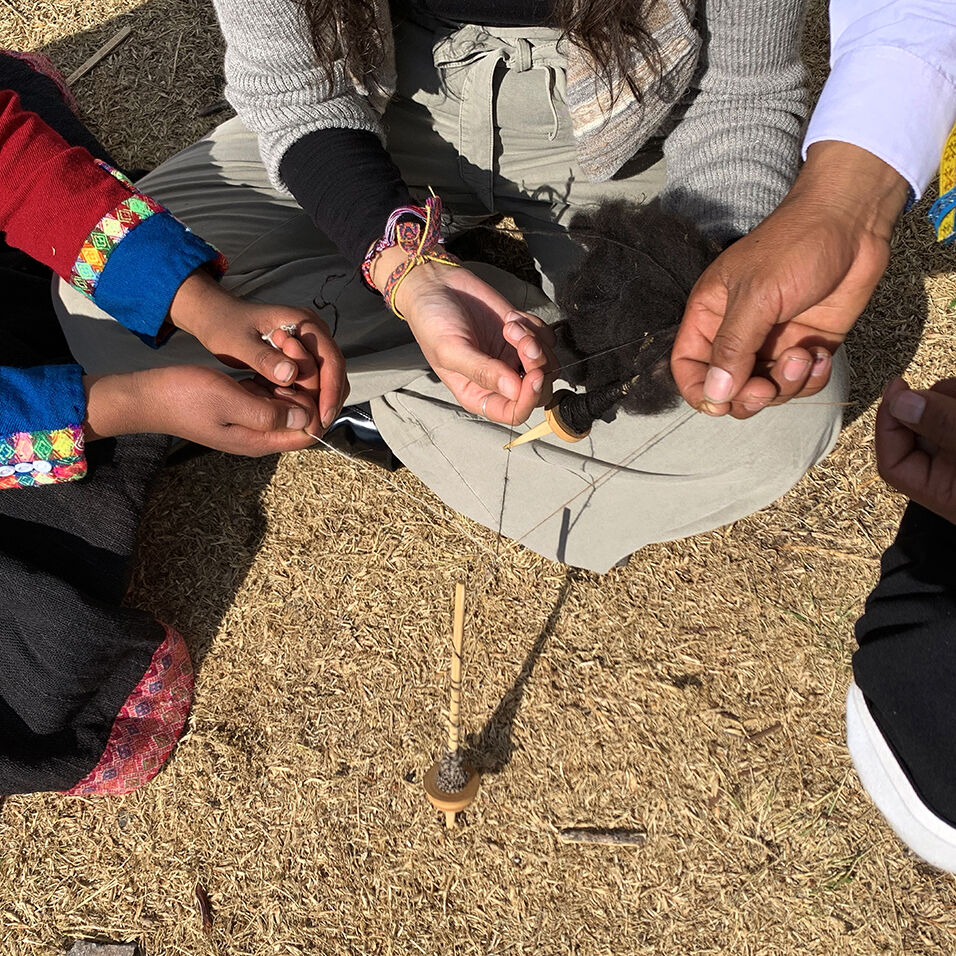 We see three sets of hands holding separate threads that are spun together on a single drop spindle on the ground between them.. 