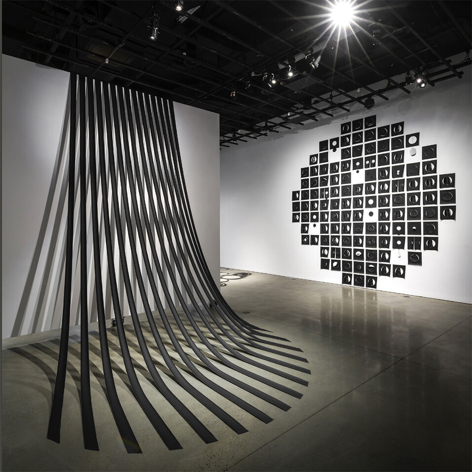 Installation shot of two monochrome artworks. To the left are ten strips of black fabric fanning out from the top of the wall to the floor. On the right wall: a diamond-shaped grid of cut black paper with empty spaces.