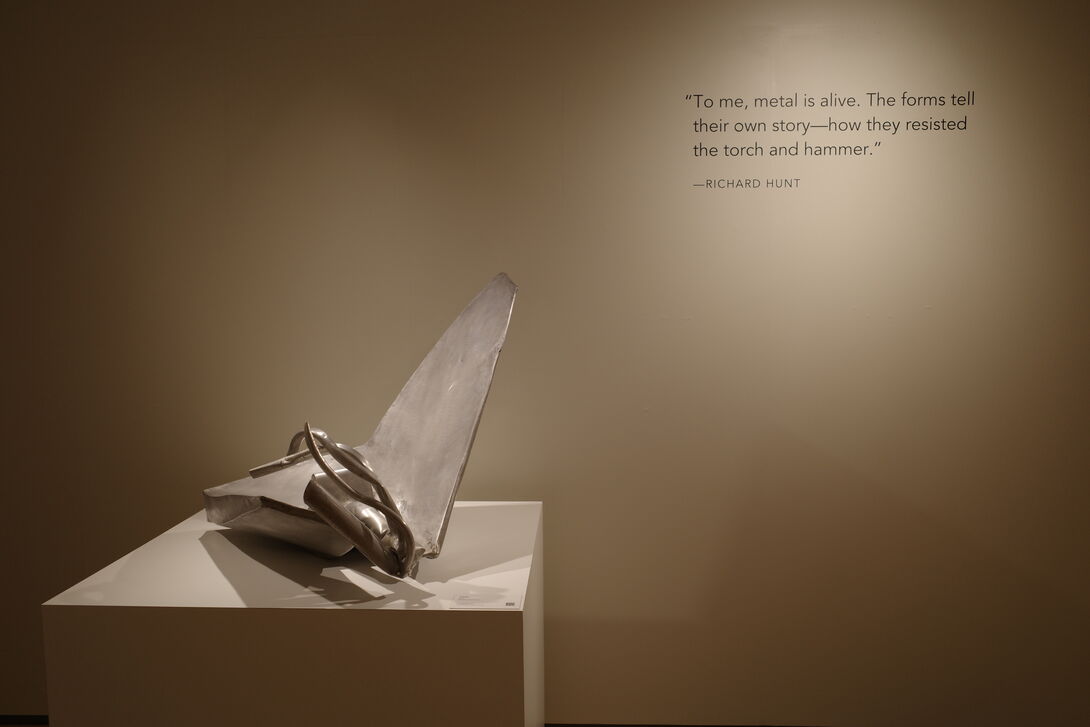 Silver abstract metal sculpture displayed on a white pedestal in a museum gallery. On the wall behind it is a quote by Richard Hunt that reads, “To me, metal is alive. The forms tell their own story—how they resisted the torch and hammer.”