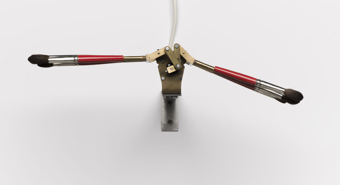 A small kinetic sculpture featuring two red-handled paintbrushes attached to a brass and metal mechanical joint mounted on a wall. The brushes extend outward like wings, connected by a central hinge mechanism with visible screws and a cord extending upward, set against a plain white background.