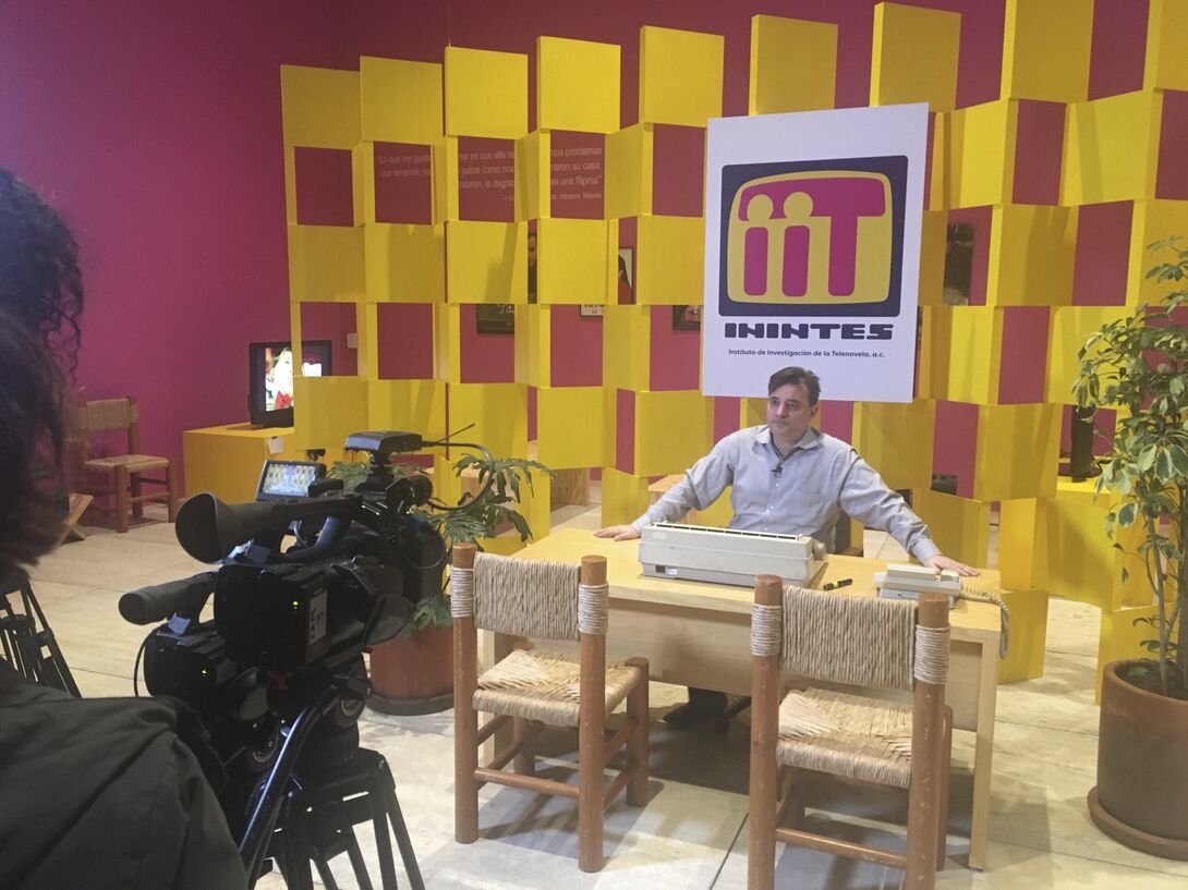 An artist sits at a wooden desk with a typewriter in a colorful installation resembling an office. Behind him, a large sign reads “ININTES” (Instituto de Investigación de la Telenovela, a.c.), displayed against a bright yellow checkered wall. The room features pink walls, potted plants, and woven chairs. A video camera films the scene from the foreground.