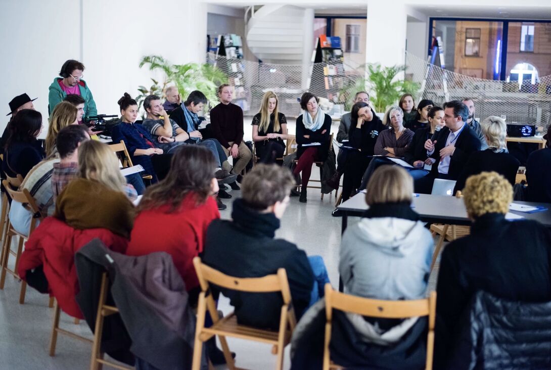 A group of about 25 people sit in a circle of wooden chairs in a bright, modern room during a discussion or workshop. One person holds a microphone while speaking, and others listen attentively, some taking notes. Tables, plants, and large windows are visible in the background.