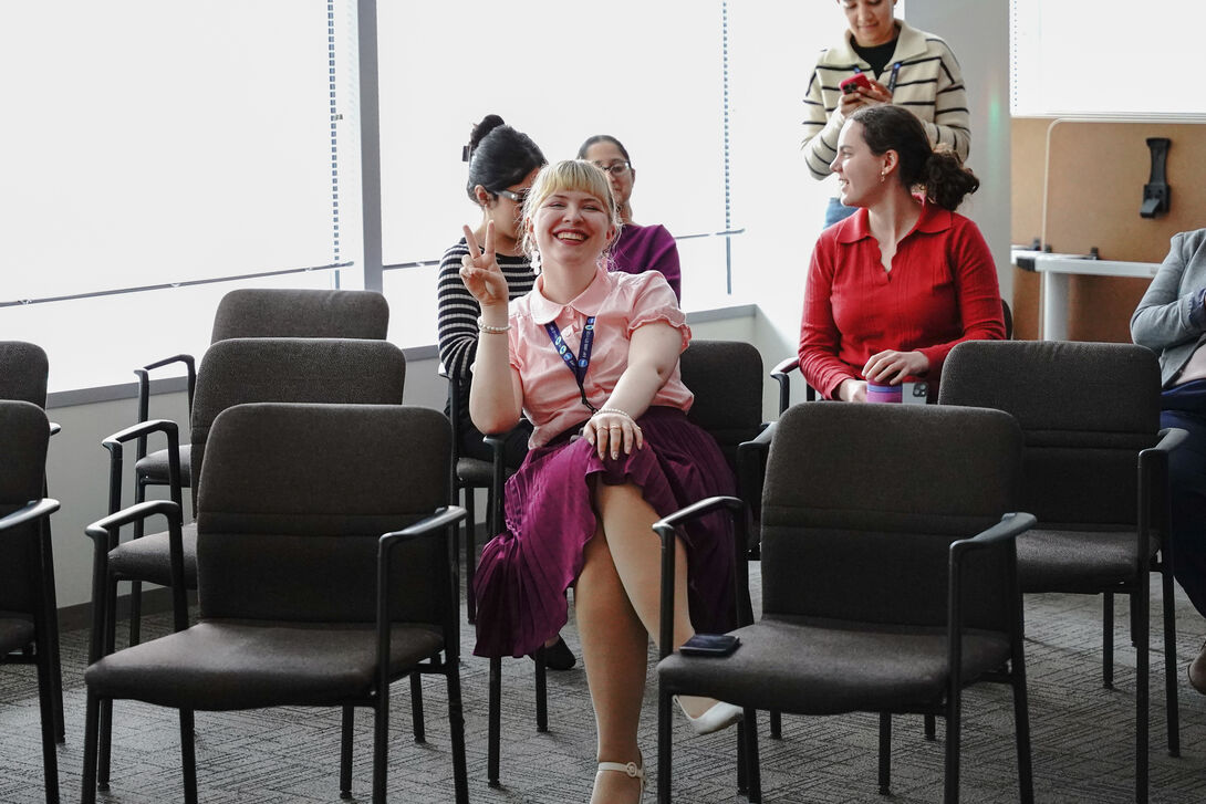 Lauren Nickels sits in a meeting room smiling and holding up a peace sign, surrounded by coworkers