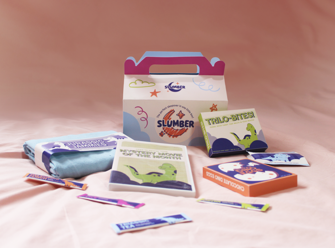 A children’s sleepover kit arranged on a soft pink backdrop, including a branded box, dinosaur-themed candy, activity cards, and folded pajamas labeled Jurassic Jammies.