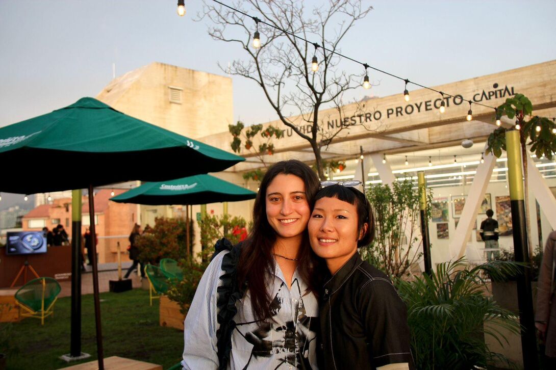 Shu stands with another artist at an outdoor evening event at the Voltaje Art and Technology Salon in Bogotá, surrounded by exhibition structures, plants, and string lights.