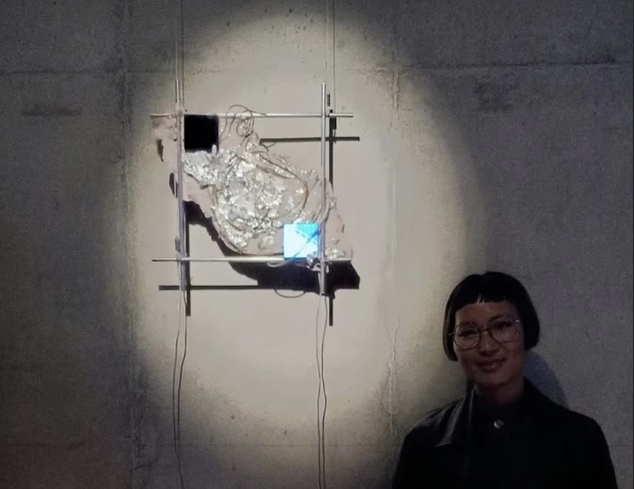 Shu stands beside a wall-mounted installation made of metal rods, concrete, resin, cables, and two small LCD screens displaying blue-toned imagery, illuminated against a textured wall at the Voltaje Art and Technology Salon in Bogotá.
