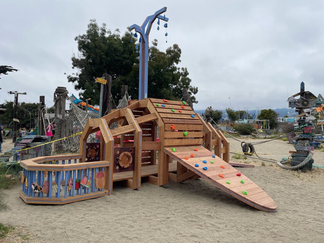 A wooden play structure in the shape of a whale sits on sand at an outdoor playground. 