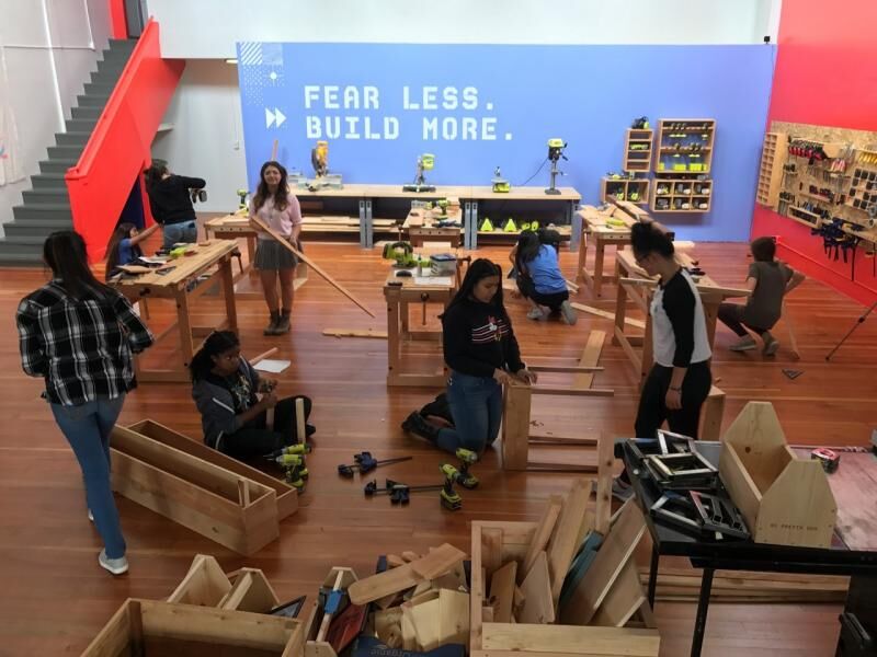 A group of young people work on wooden construction projects across a large open workshop space. Workbenches, power tools, and organized tool storage line the walls. A blue mural on the back wall reads "Fear Less. Build More."