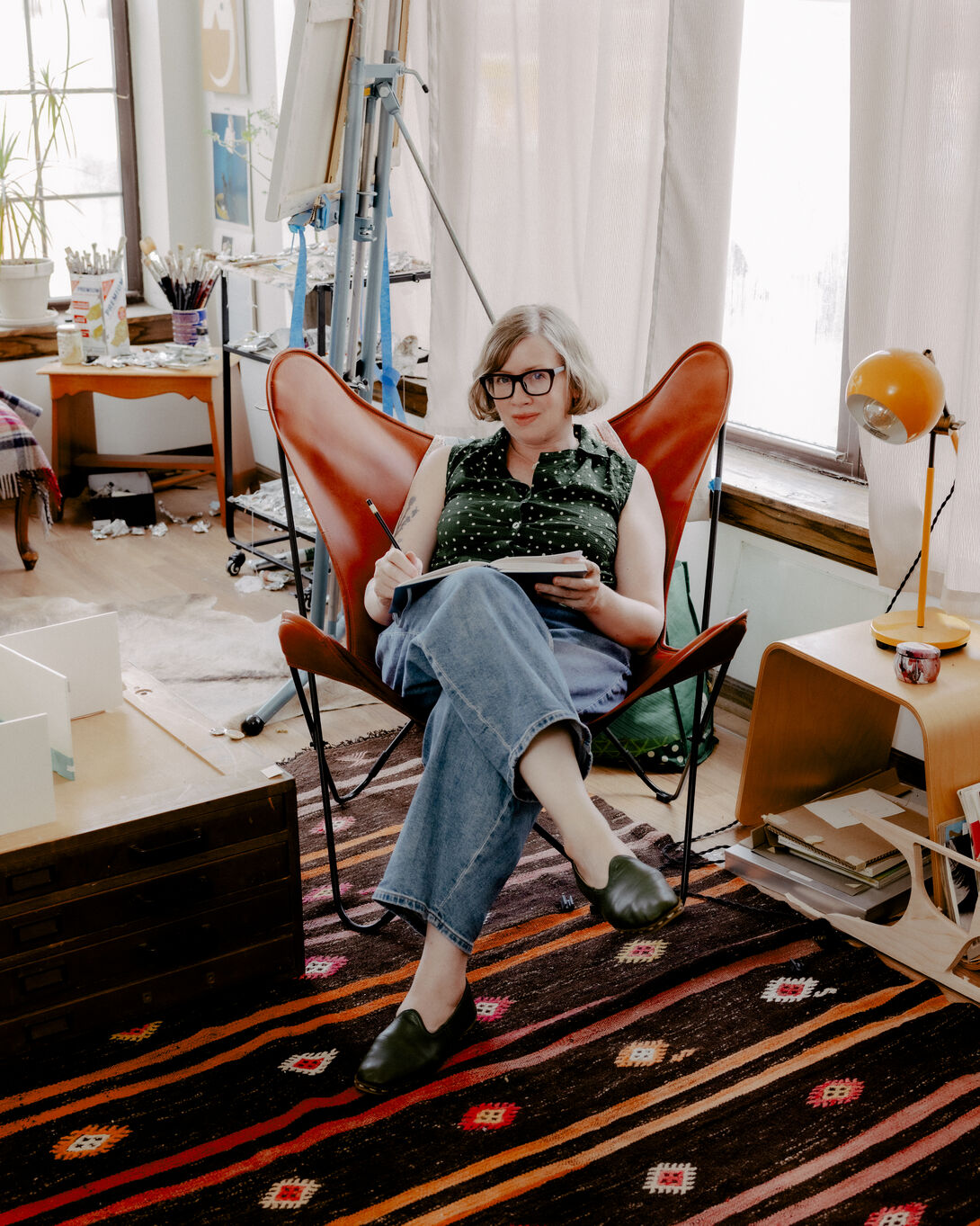 Images shows the artist, Alice Tippit, in her studio sitting in a red leather lawn chair with her right leg crossed over her left and holding a book open on her lap.