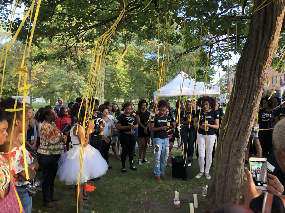 A crowd standing around a tree in a position of mourning. The tree is decorated with yellow streamers