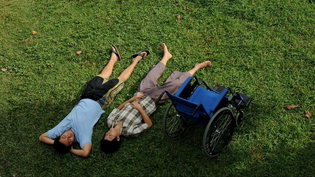 Anocha Suwichakornpong_Mundane History_2009_Courtesy of the artist and Electric Eel Films.jpeg Two men laying in the grass with a blue wheelchair next to them photographed from above.