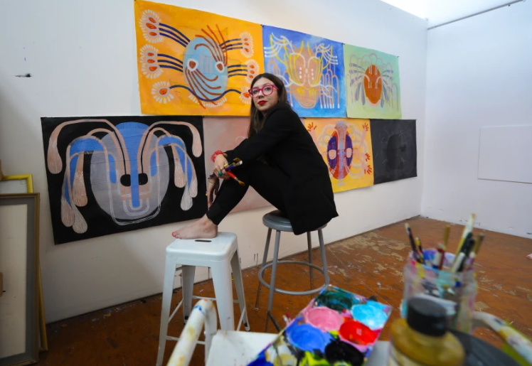 An artist sits in in her studio in front of brightly colored paintings