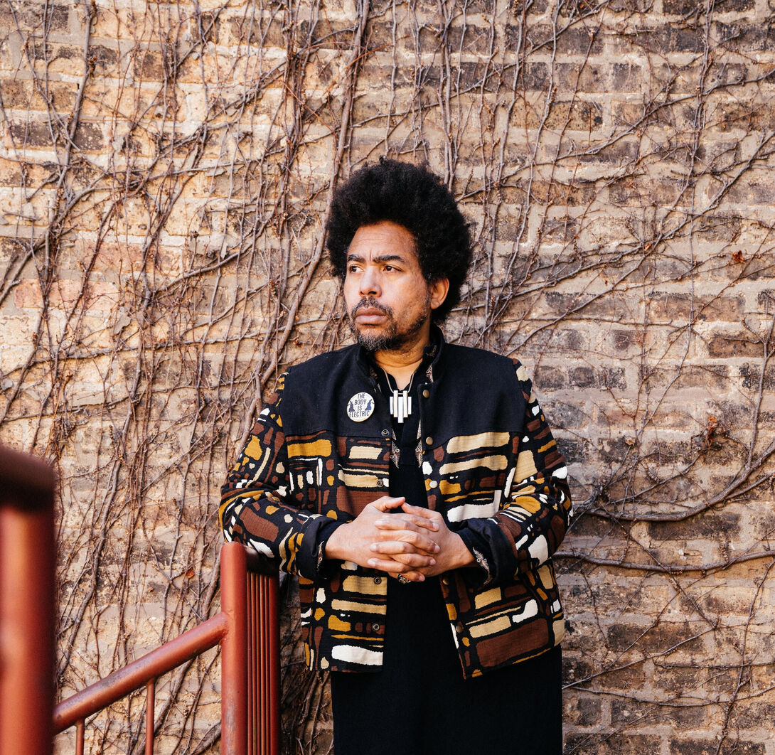 A musician stands in front of a brick wall covered in vines, looking to his right with his hands clasped in front of his chest 