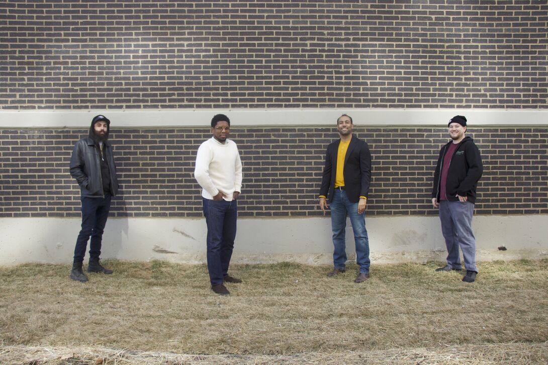 Four men stand spaced several feet apart in front of a brick wall