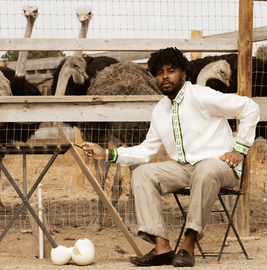 A young man sits on a chair in front of an ostrich pen 