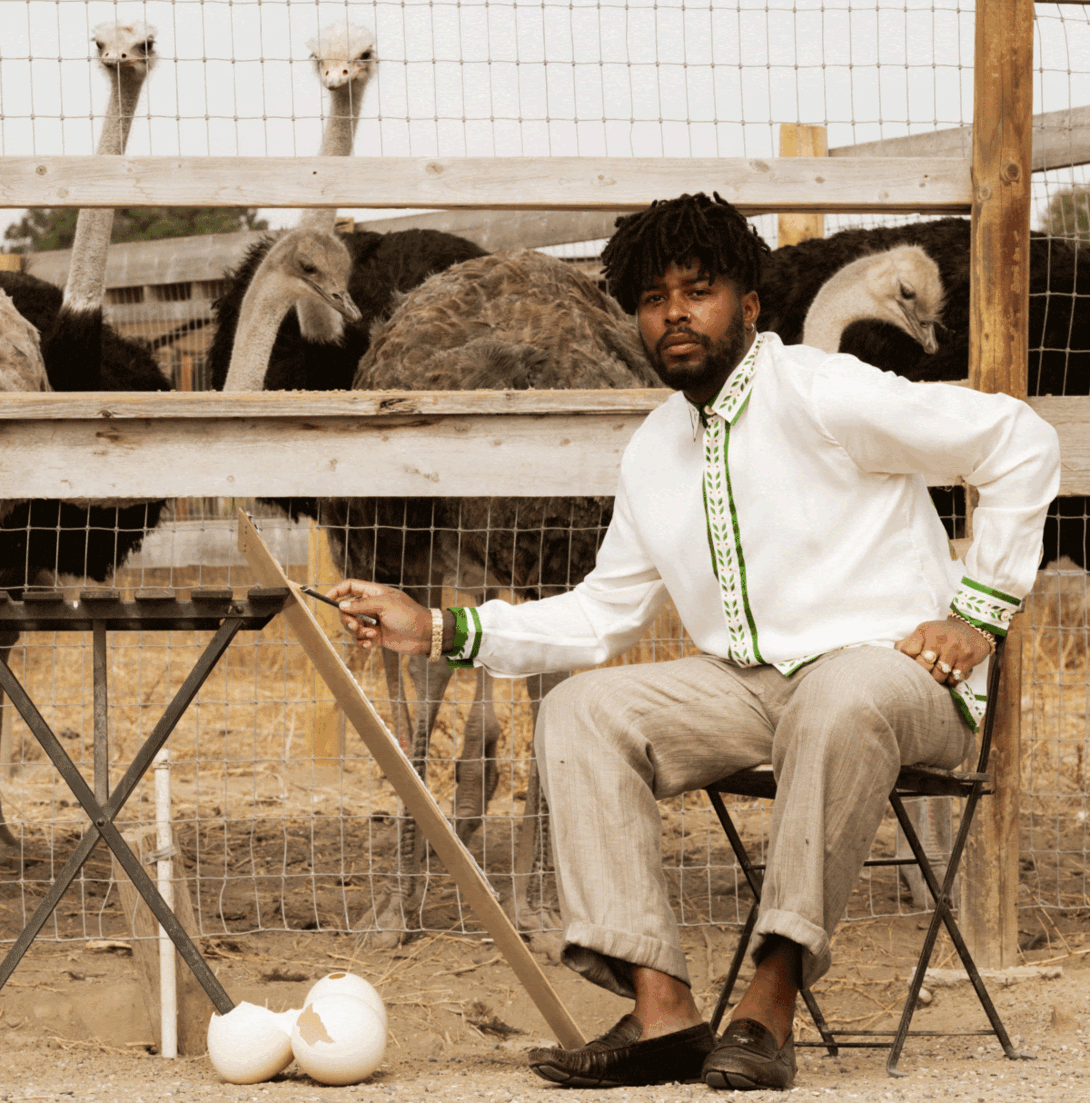 A young man sits on a chair in front of an ostrich pen